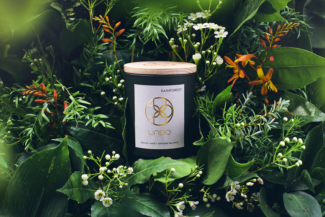 The image shows a dark green glass candle with a grey label. The label reads Rainforest at the top right. In the middle of the label is a gold brandmark, this spells the letters UNDO in a circular pattern. Under this is the name UNDO and the tagline REDUCE IMPACT. RESTORE balance.. Surrounding the candle are green leaves and orange and white flowers.